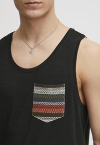 Top negru fără mâneci, cu un buzunar colorat având un model geometric în nuanțe de verde, albastru, roșu și alb. Colier argintiu cu un pandantiv.
