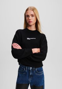 Fekete pulóver kerek nyakkal és ejtett vállakkal, fehér "Karl Lagerfeld Jeans" logóval. Magas derekú kék farmernadrággal párosítva.