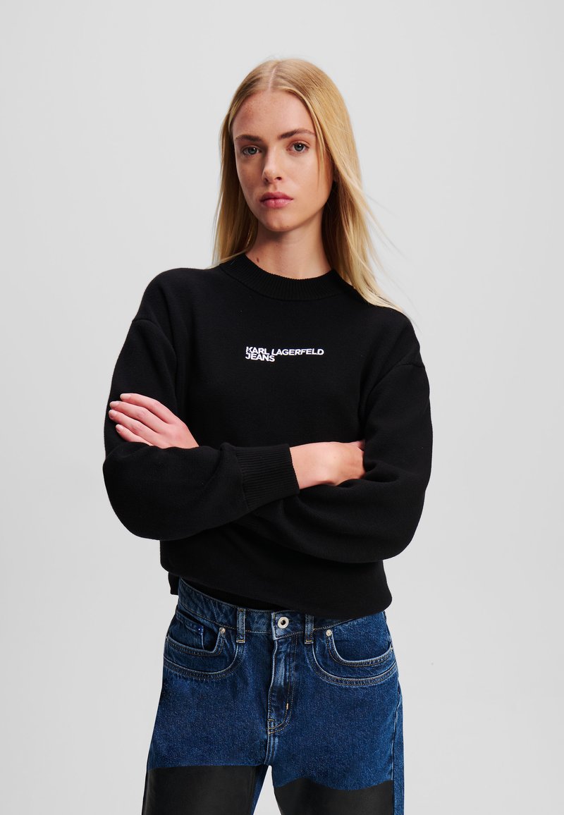 Fekete pulóver kerek nyakkal és ejtett vállakkal, fehér "Karl Lagerfeld Jeans" logóval. Magas derekú kék farmernadrággal párosítva.