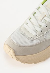 Világosszürke sneaker, amely hálós és velúr anyagok keverékéből készült, fehér cipőfűzőkkel és egy ferde mintázatú, textúrált bézs talppal rendelkezik.