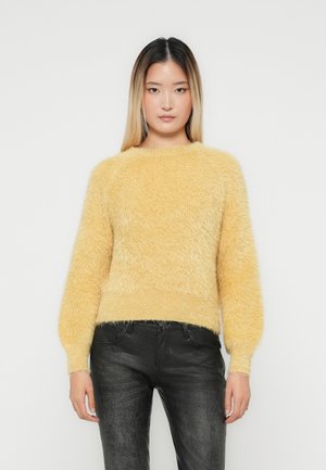 Vrouw staat rechtop, kijkt naar voren en draagt een fuzzy gele trui met pofmouwen en zwarte gestructureerde broek tegen een plain witte achtergrond.