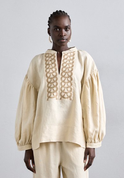 Femme avec des cheveux tressés portant un blouse ample crème avec un panneau avant brodé et un pantalon large assorti sur fond uni.