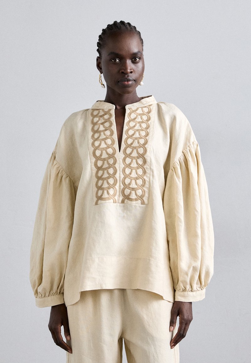 Femme avec des cheveux tressés portant un blouse ample crème avec un panneau avant brodé et un pantalon large assorti sur fond uni.