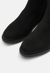 Botines de ante negros con punta redonda, que presentan una cremallera, detalles cosidos a lo largo de la suela y una textura mate suave.