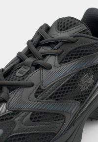 Gros plan d'une chaussure de sport noire avec des panneaux en mesh, des empiècements synthétiques, des lacets noirs et un petit logo de crocodile sur le côté.