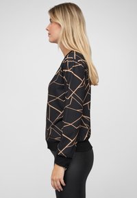 Eine Frau mit langen blonden Haaren, die einen schwarzen Sweatshirt mit beigen geometrischen Linien und schwarze, eng anliegende Hose trägt, steht im Profil vor einem grauen Hintergrund.