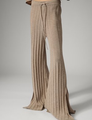 Beżowe prążkowane spodnie dzianinowe z szerokimi, rozkloszowanymi nogawkami, talią z troczkiem i małymi bocznymi rozcięciami na dole, noszone przez stojącą osobę.