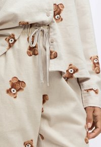 Beige lounge set met teddybeerprint. Zacht materiaal, ontspannen pasvorm. Voorzien van knopen, trekkoorden en zichtbare stikseldetails.