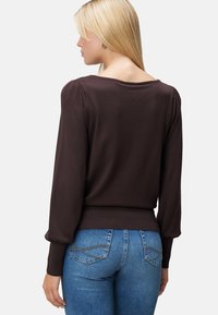 Femme blonde de dos portant un haut marron foncé à manches longues avec des épaules bouffantes et un jean ajusté bleu avec des poches arrières cousues.