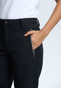 Sorte bukser har en lynlåslomme på låret, lavet af et glat stof. Livremmen inkluderer en knap og bæltesløjfer.