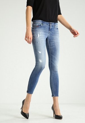 Femme portant un jean skinny bleu usé avec fermetures éclair aux chevilles et talons hauts noirs, crop top noir visible au-dessus de la taille.