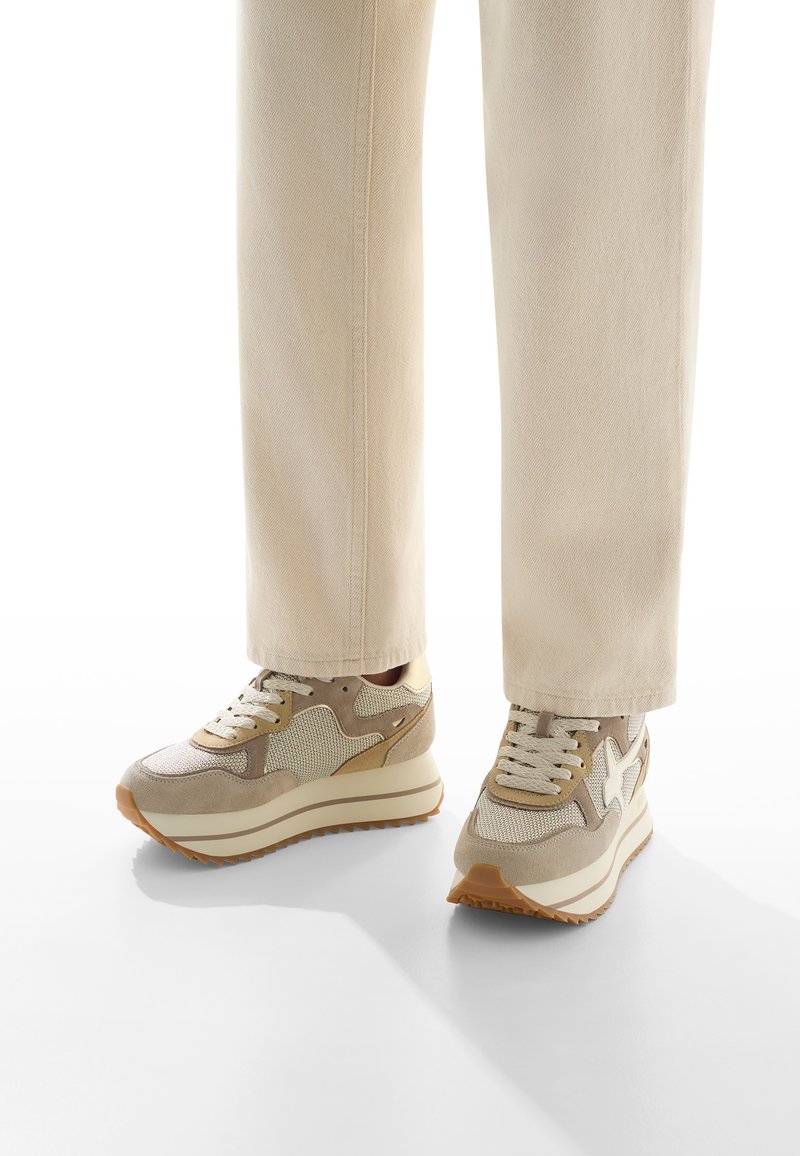 Persona che indossa sneaker beige con suole bianche e pantaloni beige chiaro su uno sfondo bianco, piedi leggermente divaricati e che proiettano ombre.