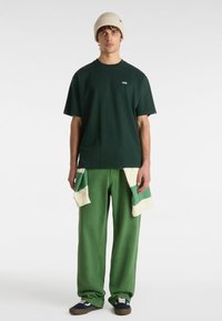 Green cotton t-shirt with short sleeves and Vans logo, paired with wide green pants featuring green and cream panel accents, finished with blue sneakers.