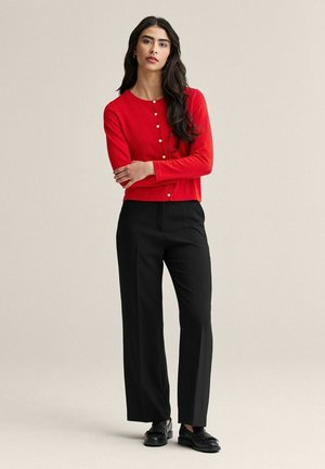 Kvinde med langt mørkt hår iført en knappet rød cardigan, sorte vidde bukser og sorte loafers, stående med armene krydsede.