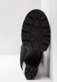 Botín negro hasta el tobillo con un upper de tejido texturizado y una suela de goma resistente. La suela presenta un patrón de tracción distintivo.