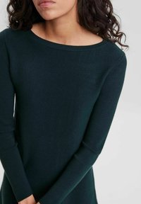 Femme aux cheveux bouclés et foncés portant un haut en tricot ajusté à manches longues de couleur vert foncé avec un col rond, contre un fond uni.