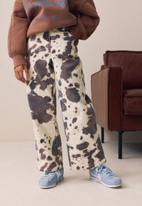 Pantalon en denim à jambes larges, imprimé vache en beige avec des éclaboussures noires, assorti à des baskets bleu clair au design texturé.