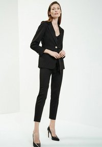 Czarny garnitur z dopasowaną marynarką z zapięciem na złoty guzik i slim fit spodniami, w połączeniu z czarnymi szpilkami. Gładka tekstura materiału.