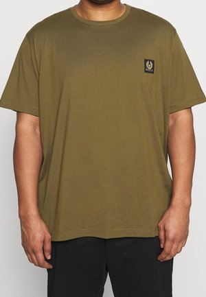 Mand iført olivengrøn kortærmet t-shirt med lille emblem på venstre bryst og sorte bukser, mod en ensfarvet baggrund.