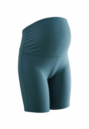Personne enceinte portant un short de sport de maternité bleu sarcelle et un haut court assorti, debout contre un mur de briques blanches.