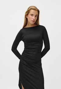 Μαύρο φόρεμα με μακριά μανίκια, εφαρμοστή γραμμή, ριγέ υφή και ρουστίκ πλευρές. Περιλαμβάνει λαιμόκοψη μπάτζα και σκίσιμο στο µηρό από τη μία πλευρά.