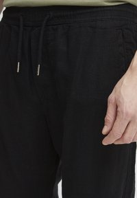 Pantalons en lin noir avec une taille élastique, fermeture à cordon, et poches latérales. Le tissu a une finition texturée.