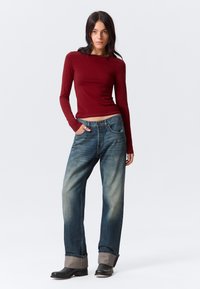Frau steht mit einer Hand in der Tasche, trägt ein enges, kastanienbraunes Langarmoberteil, lockere blaue Jeans mit hochgerollten Säumen und schwarze Stiefel.