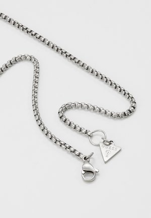 Collier chaîne boîte en argent avec fermoir mousqueton et pendentif triangulaire au logo Guess sur fond blanc uni.