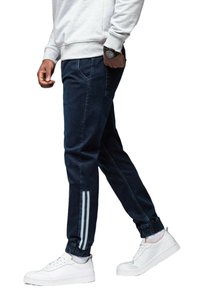 Pantalones jogger de mezclilla oscura con un corte ajustado, que cuentan con acentos en rayas gris claro en la parte baja de las piernas y puños elásticos. Se llevan con zapatillas blancas.