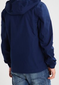 Giacca con cappuccio blu navy con polsini elasticizzati e texture liscia, caratterizzata da un design semplice senza loghi o motivi visibili.