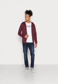 Maronfarvet zip-up hoodie over en hvid t-shirt med rød skrift, parret med blå jeans og grå sneakers. Enkel, afslappet design.