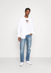 Witte button-up shirt over een grafisch t-shirt, gecombineerd met lichtblauwe jeans en witte sneakers. Eenvoudig ontwerp met een relaxte pasvorm.