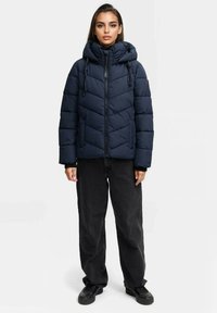 Granatowa kurtka puchowa z kapturem, pikowana, z zamkiem błyskawicznym z przodu i dwoma bocznymi kieszeniami, w połączeniu z czarnymi spodniami i czarnymi butami wsuwanymi.