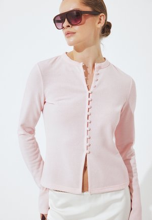 Donna che indossa un cardigan rosa chiaro abbottonato con bottoni raccolti, gonna bianca e occhiali da sole marroni oversize, capelli raccolti in uno chignon basso.