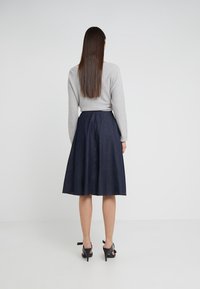 Šedý svetr v kombinaci s sukní ve tvaru A délky po kolena v námořnické modři, která má hladkou texturu a rozšířený design, doplněná černými sandály na podpatku.