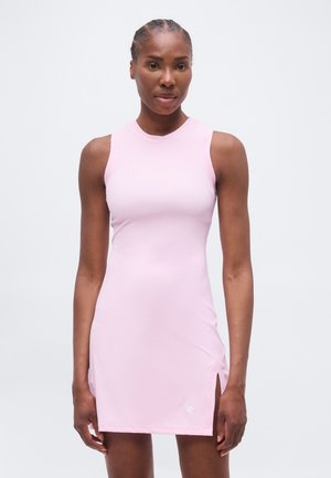 Γυναίκα με πλεξούδες, φορώντας ένα αμάνικο, ανοιχτό ροζ φόρεμα μέχρι το γόνατο με μια μικρή πλευρική σχισμή, στέκεται μπροστά σε απλό φόντο.