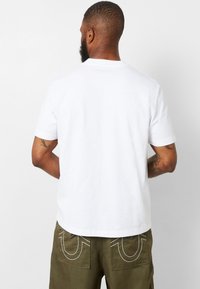 Camiseta blanca de algodón con mangas cortas y cuello redondo, vista desde la parte de atrás. Sin patrones ni logotipos visibles; textura suave.