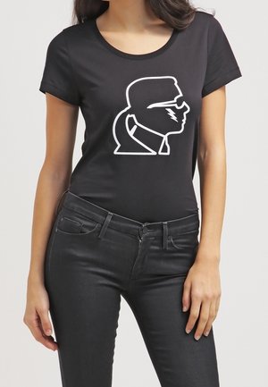 Frau trägt ein schwarzes, eng anliegendes T-Shirt mit einer weißen Grafik eines Personenprofils mit Sonnenbrille und Pferdeschwanz.
