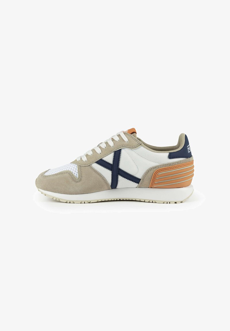 Zapatilla deportiva en ante beige y malla blanca, con acentos azules, un diseño de talón texturizado y una suela de goma blanca con patrones de relieve.