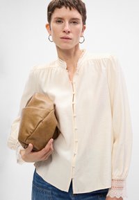 Blusa color crema con colletto alto, pattina di bottoni e polsini con pizzo. Abbinata a una borsa a tracolla in pelle marrone.