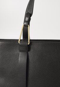 Black leather tote bag with textured surface, featuring a gold-tone metal hardware accent and adjustable strap near the opening.