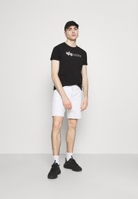 Juodas medvilninis marškinėliai su "Alpha Industries" logotipu, derinami su baltais džinsiniais šortais. Modelis vilki juodus sportbaigus ir juodą kepurę.