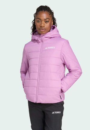A woman with curly hair wears a hooded lavender jacket and black pants, posing confidently against a light background.