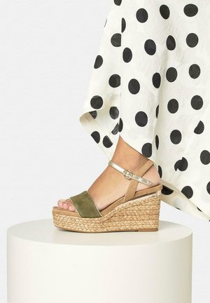 Pie con una sandalia de cuña con pulsera en el tobillo en colores oliva y beige, con plataforma tejida, junto a una falda blanca con grandes lunares negros.