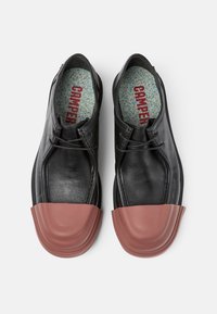 Zapatos de cuero negro con una puntera redondeada de goma marrón, con cordones planos y una plantilla moteada. El diseño combina elegancia y durabilidad.