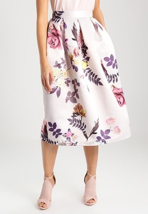 Kvinde iført en hvid midi-nederdel med lilla, pink og gul blomsterprint og plisseringer, kombineret med lyserøde åbne hæle.