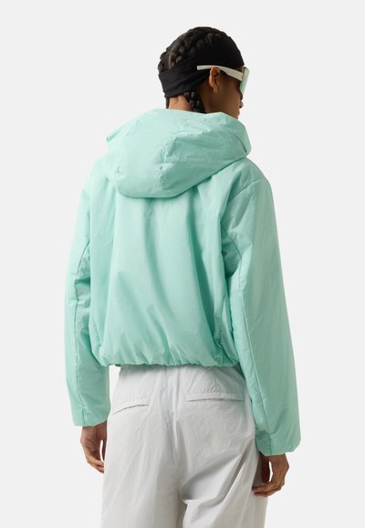 Person with braided hair wearing mint green hooded jacket, white pants, black headband, and white-framed sunglasses, shown from the back.