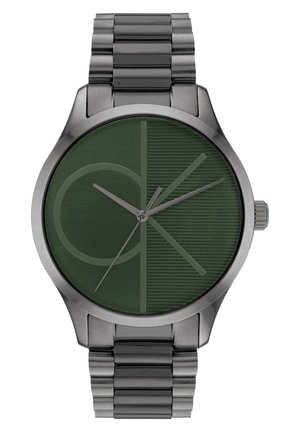 Montre à cadran vert avec une surface texturée, arborant un audacieux logo "CK". Bracelet en métal argenté brillant, boîtier rond avec des aiguilles minimalistes.