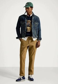 Ung man iklädd en grön keps, denimjacka, tröja med teddy-björn, khakifranska byxor och blå sneakers som står mot en enfärgad bakgrund.