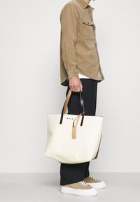 Bolso tote grande de color blanco con detalles en cuero negro y marrón claro, con forma rectangular y una etiqueta colgante. El diseño es minimalista.